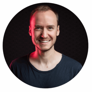 Porträtfoto eines lächelnden Mannes mit schwarzem T-Shirt und roter Lichtkante vor dunklem Hintergrund